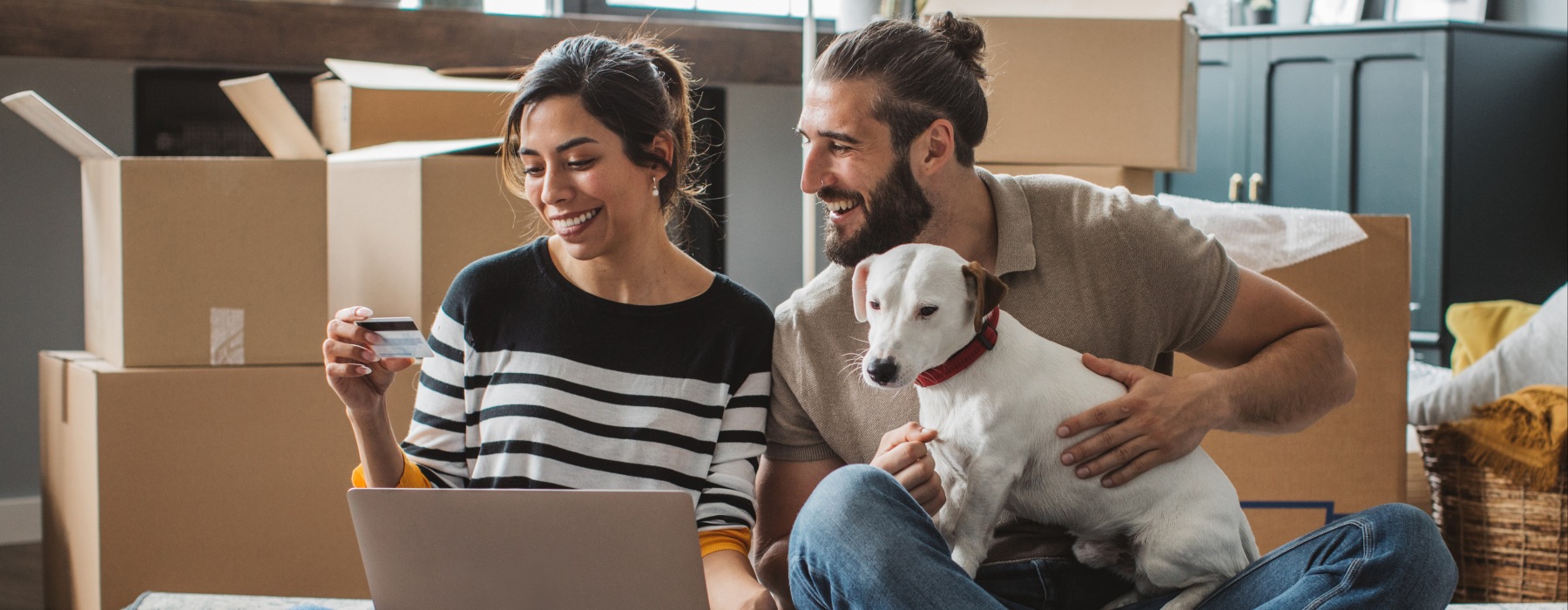A smiling couple sitting on the floor of their new pet-friendly apartment surrounded by moving boxes, using a laptop and credit card with their dog.