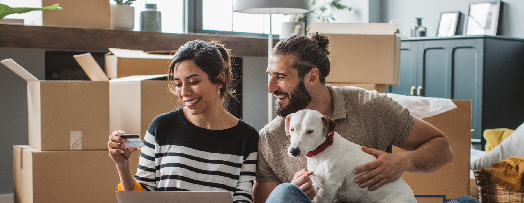 A smiling couple sitting on the floor of their new pet-friendly apartment surrounded by moving boxes, using a laptop and credit card with their dog.